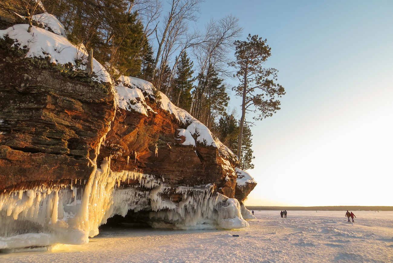 Photograph of sandstone cliffs draped in icicles and people walking across a frozen lake.