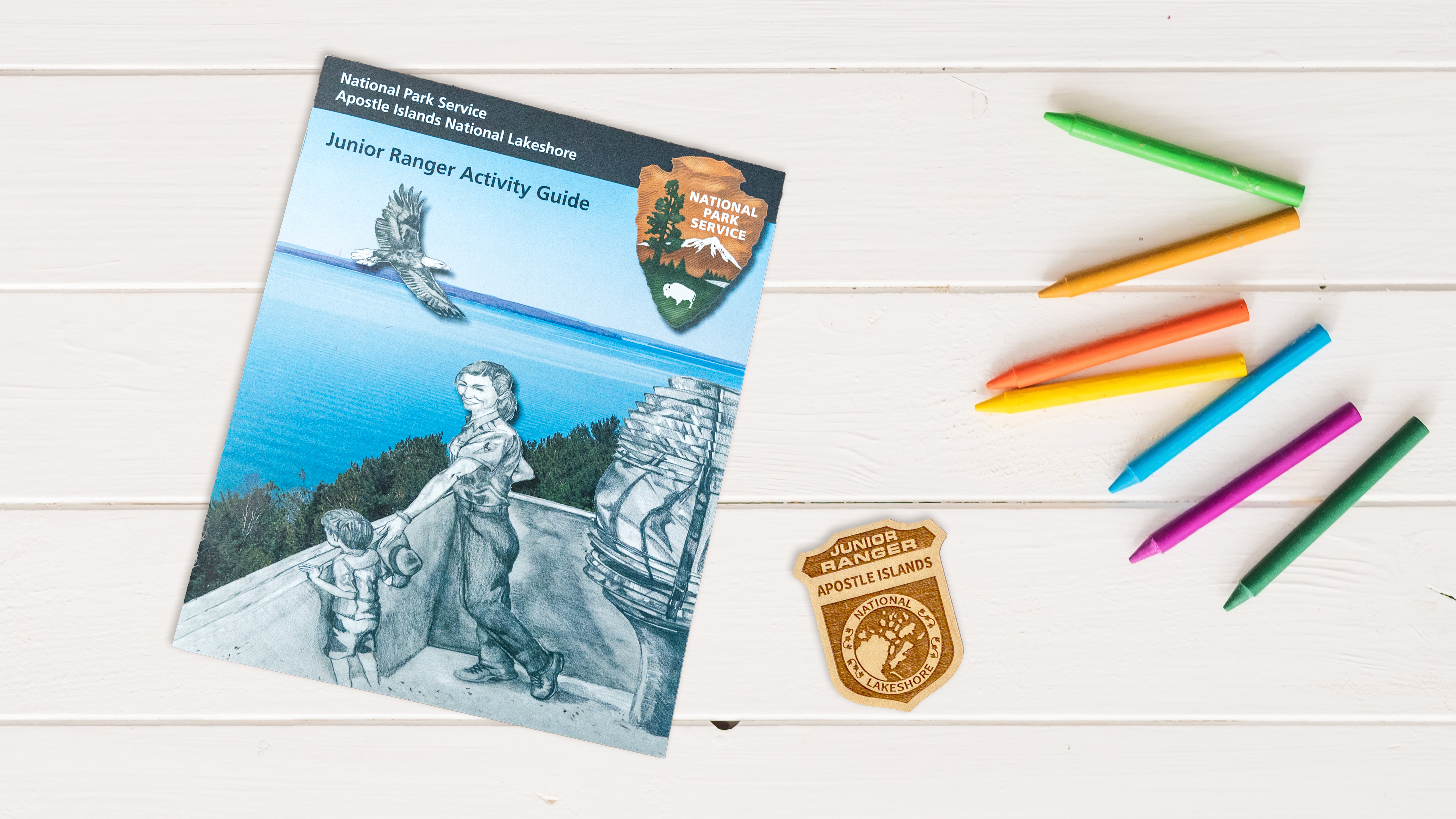 Photograph of booklet, crayons, and Apostle Islands Junior Ranger badge on a white table/