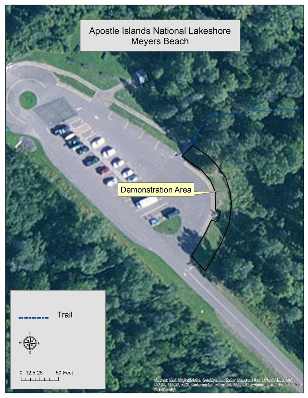 Aerial view of a paved parking lot with cars surrounded by trees. An area in the grass, on the eastern edge of the lot, is outlined in black and designates the first amendment demonstration area for the National Lakeshore.