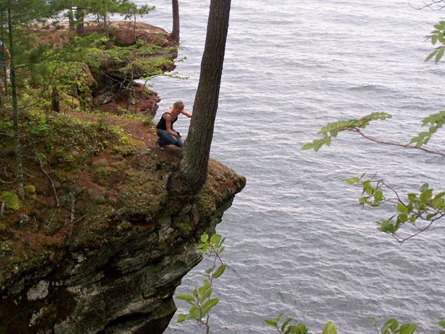 A kneeling woman at the edge of a cliff over the water, leaning against a tree trunk.