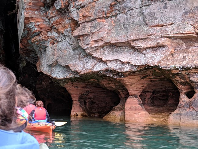 View of kayakers from the back, entering caves in a sandstone formation.