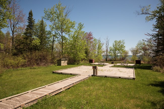 Photograph of a grassy area with wooden walkway leading to a picnic tables on platforms. Trees and a lake are visible in the background on a clear, sunny day.