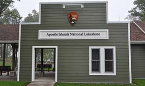 Front of a building with grey siding, an NPS Arrowhead, and Little Sand Bay Visitor Center.
