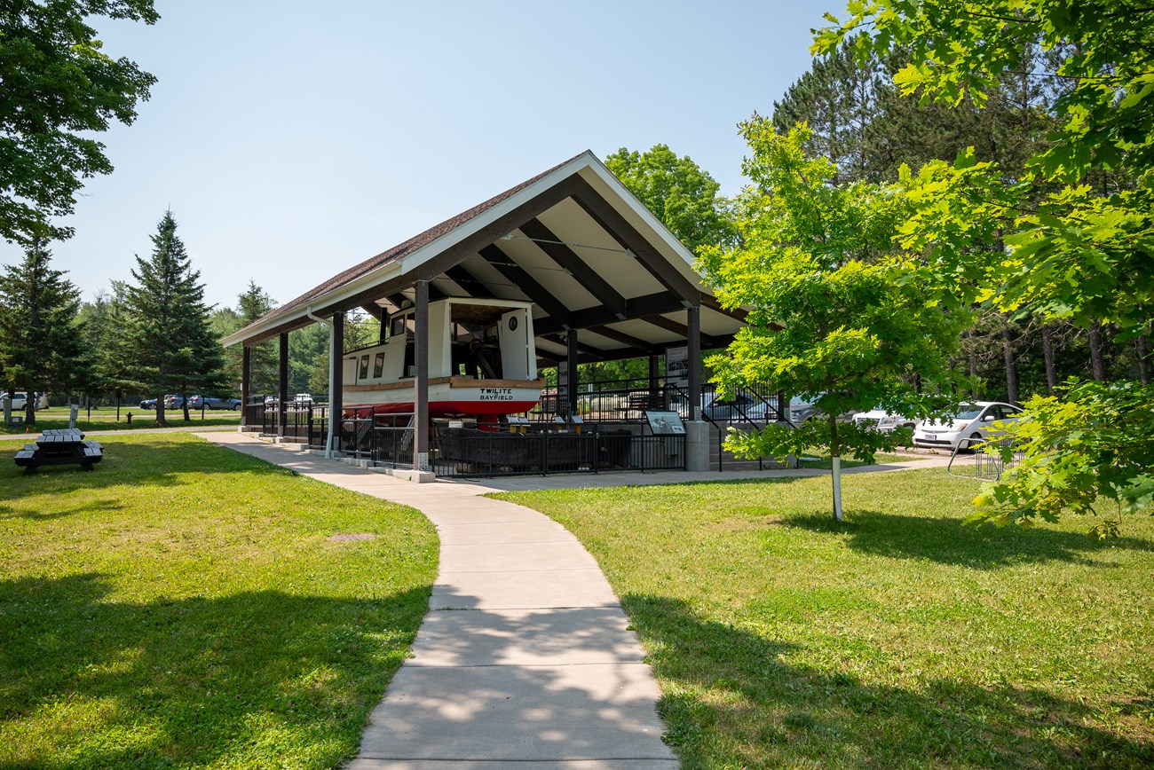 Photograph of cement walkway leading towards a large pavilion. Beneath the pavilion is a historic fishing boat with "Twilite of Bayfield" written in black text on the stern.