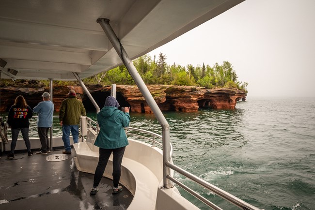 Passengers look out from the back of a passenger boat at sandstone caves under a cloudy sky.