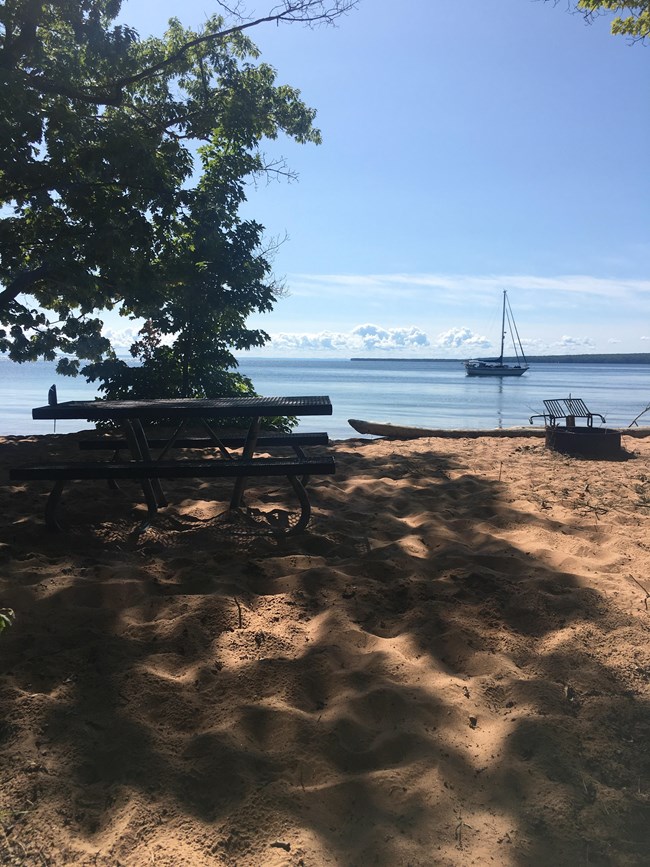 A shaded picnic table in the sand looking out at a lake with an anchored sailboat.