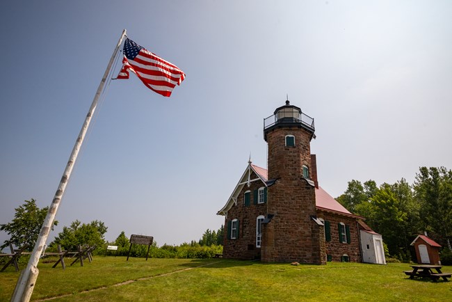 Photograph of a brick lighthouse under a blue sky with an American flag fluttering in the foreground.