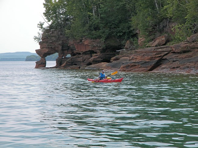 A kayak paddler near sandstone arch on lake.