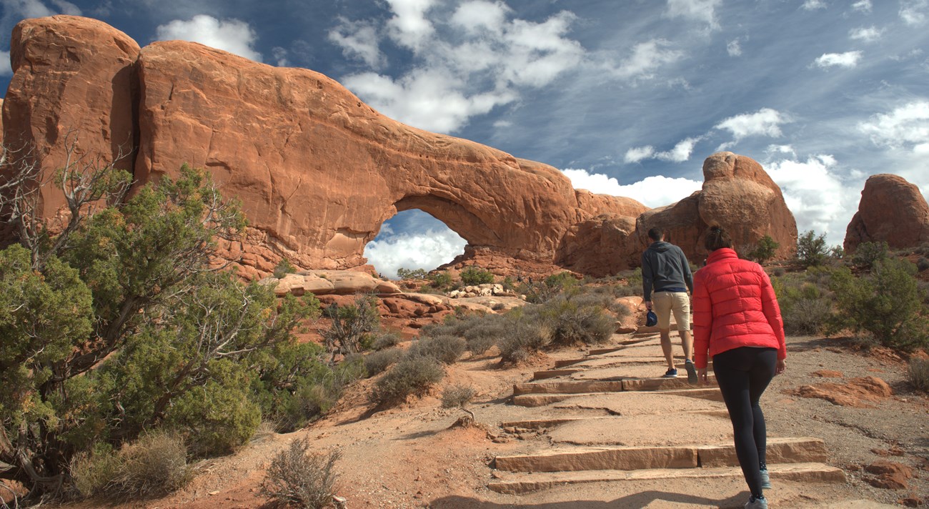 two hikers climb trail past low shrubs toward large stone arch under blue sky with clouds