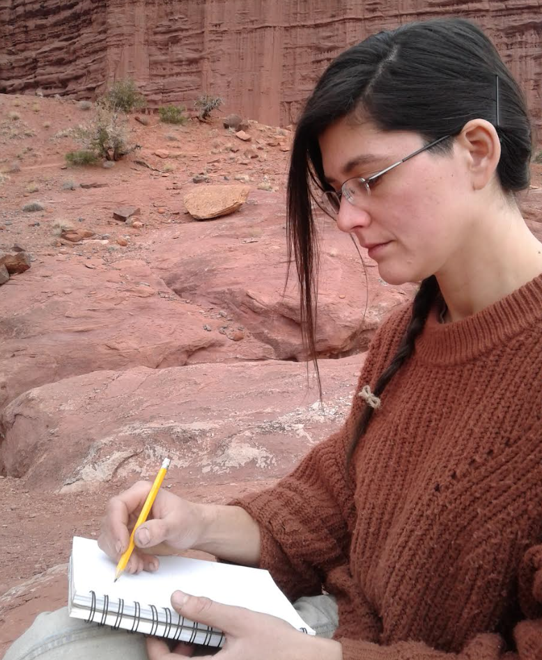 A light skinned woman with dark hair and glasses sketches in a notebook, seated near a red sandstone wall
