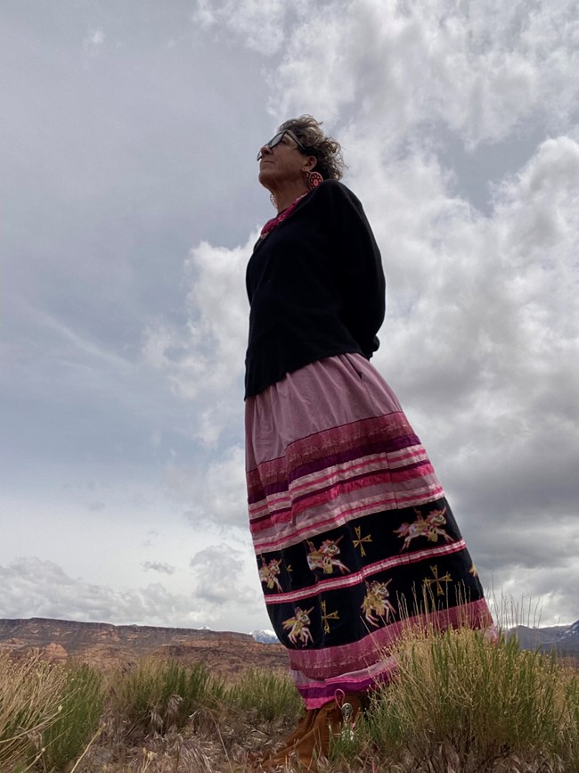 woman in colorful patterned skirt stands on dry grasses looking up at blue sky