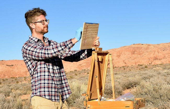 A man looking off in the distance, holds up a canvas with an easel in front of him. He is outside in a sage brush and red rock background with a bright blue sky.