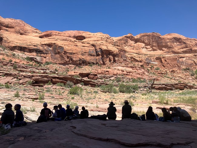 A group of adults and children sit on a long stretch of sandstone. The people are all silhouetted. A canyon wall and green vegetation are in the background.