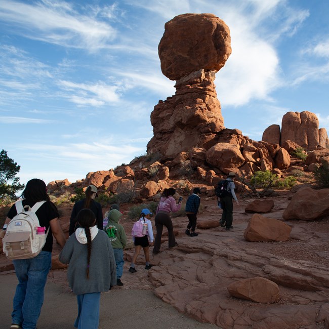 A group of students and adults follow a park ranger up uneven rock steps in front of a large sandstone formation