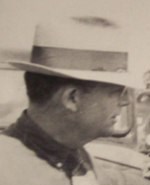 A black and white photo of the profile of a mans face as he looks at something out of frame. He is wearing a collared shirt and a fedora.