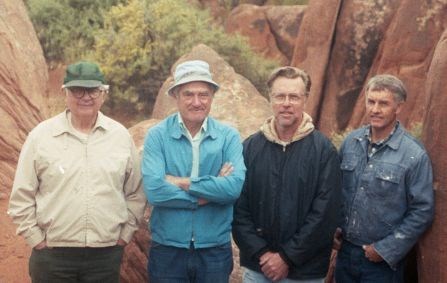 Four men stand shoulder to shoulder smiling at the camera in front of a red rock background.