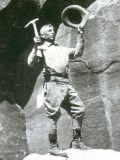 A black and white photo of a man standing on a rock ledge holding a hammer and a hat.