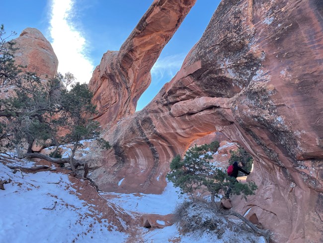 Large rock arch with two openings surrounded by snow. A person is hiking below the arch.