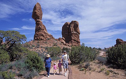 A family of four holds hands, walking on a sidewalk through a desert landscape with sand and short green trees. A large, top-heavy sandstone feature towers behind them
