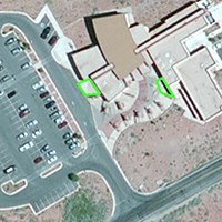 A satellite image showing a building, a parking lot, and a road. Two areas next to the building are outlined in green.