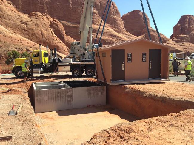 Construction workers can be seen wearing yellow vests and hats surrounding a crane lifting a pit toilet building into place over the underground waste storage tanks.