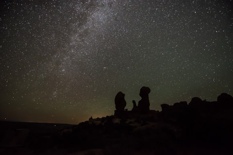 The milky way streaks across a blue and green night sky behind the silhouettes of rock spires.