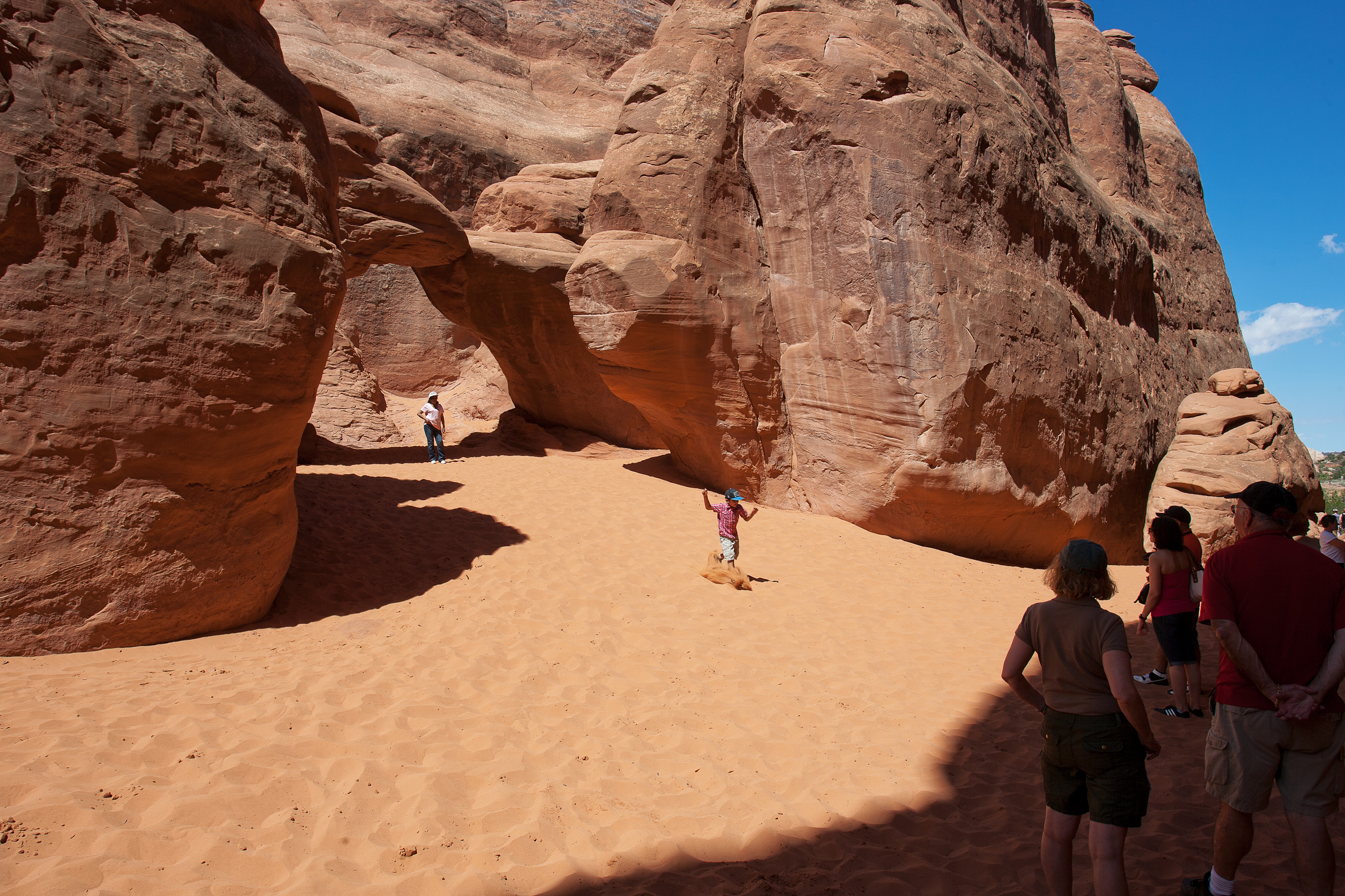 people stand in shade near and beneath sandstone arch, surrounded by sunlit loose sand