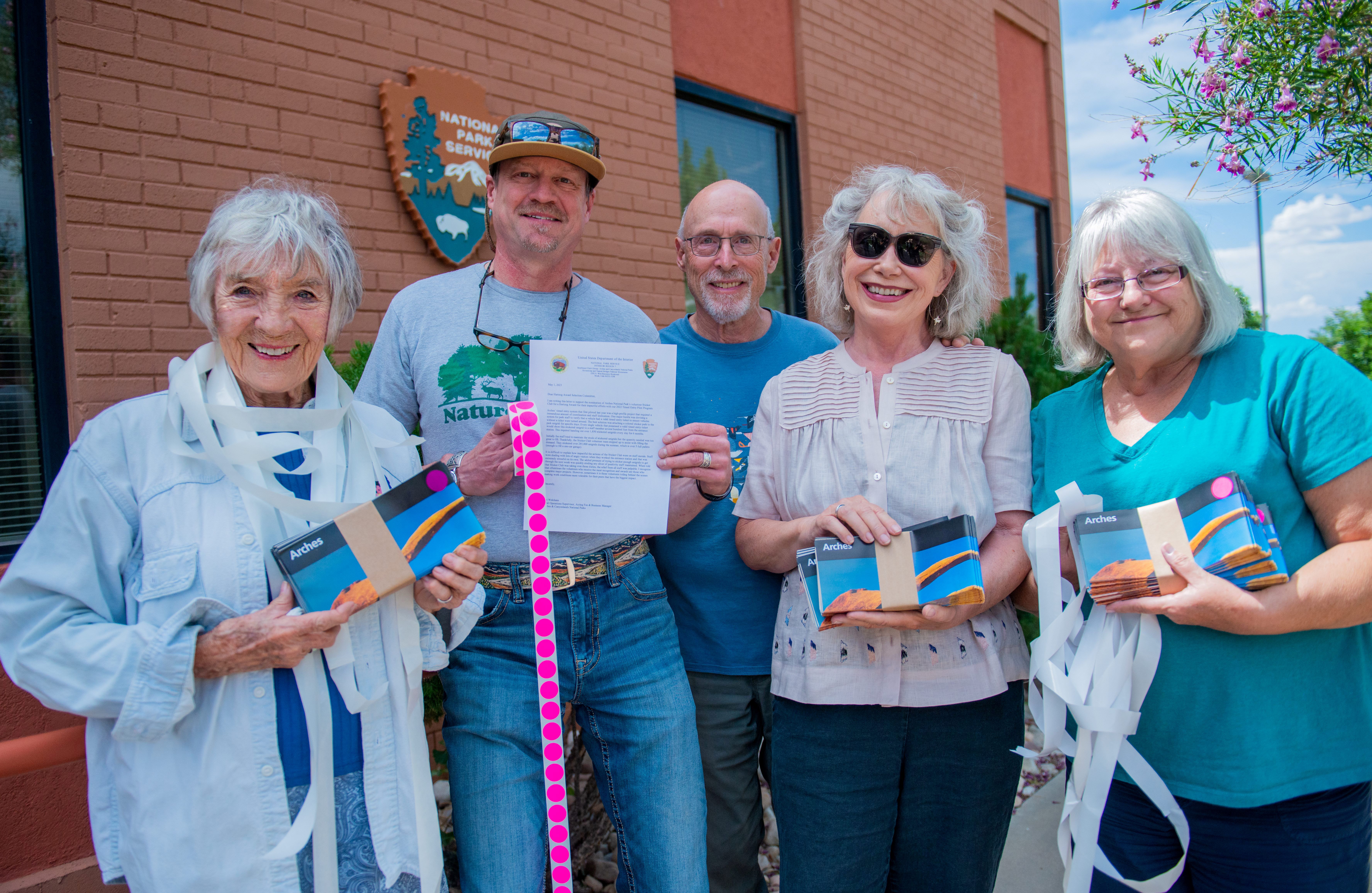 Five seniors stand outside a building, holding Arches maps and rolls of pink stickers