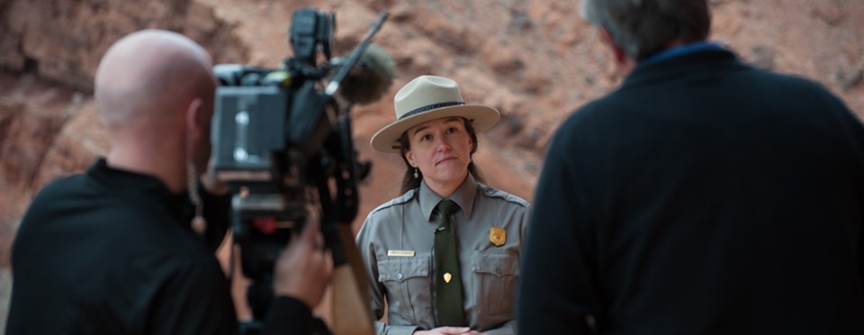 A female park ranger stands in front of a video camera surrounded by steep red rock walls.
