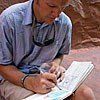 A man is seated on red rock drawing on a notebook in his lap.