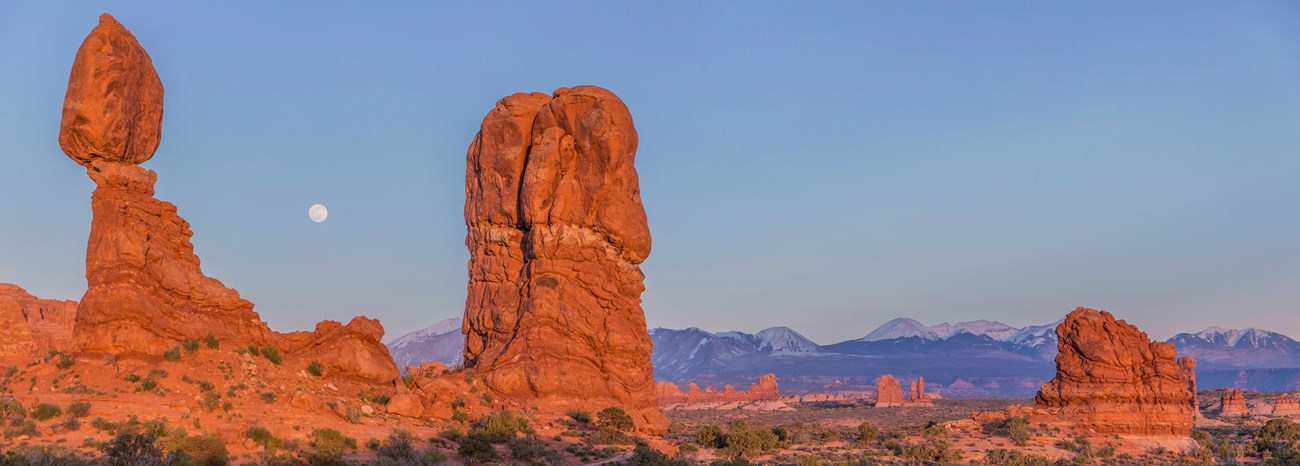 A landscape of spread out towering rock pillars. One looks like a large boulder is balancing on a pedestal. The rocks are red and are glowing from a rising sun. The sky is blue and a full moon is setting, into the distant snowcapped mountains.