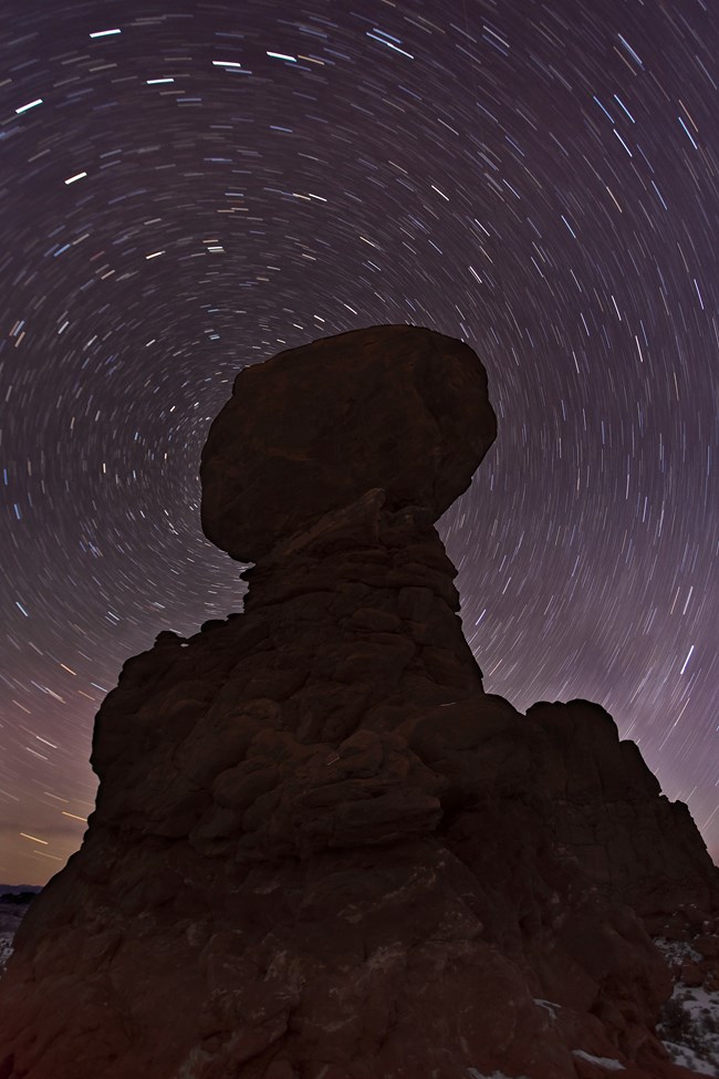 This photo was taken with a long exposure meaning the stars are small white lines spinning around a silhouetted large rock formation
