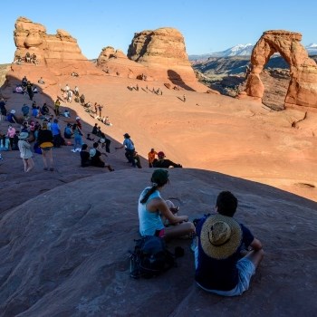 A crowd of people sit in a rock basin near a free standing natural rock arch.