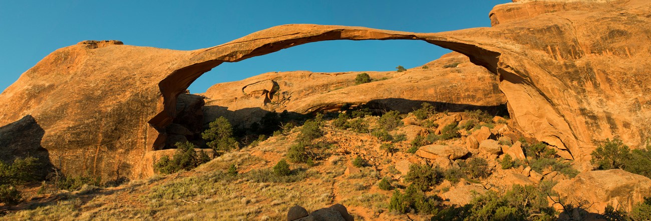 A thin piece of red rock arches across the green and sandy desert landscape, connecting two large rocks.