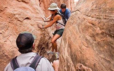 Two people navigate through a narrow gap between red rocky faces while a ranger waits.