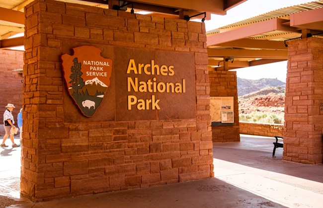 stone wall displaying large NPS arrowhead and the words "Arches National Park" with similar walls on surrounding plaza