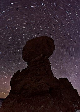 A tall pillar of rock is silhouetted by the black and deep purple night sky. The photo was taken with a long exposure, causing the stars to become bright circling lines in the sky.