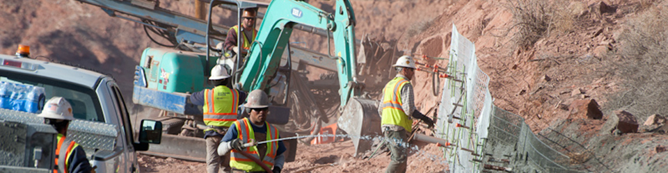 5 constructions workers in yellow safety vests and white helmets work on retaining a wall of red rock, dirt, and sand. There is a white truck and a teal excavator.