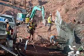 5 constructions workers in yellow safety vests and white helmets work on retaining a wall of red rock, dirt, and sand. There is a white truck and a teal excavator.