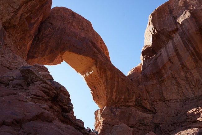 Large sandstone arch with people scrambling at the base. Clear sky through the arch.