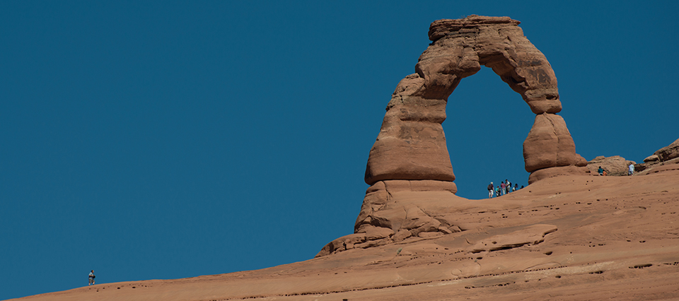 A large sloping red rock face with a unique rock formation jutting out of it. It is an arch shaped like an upside U. The blue sky can be seen through the wide opening in the rock. The people standing below it look very small.