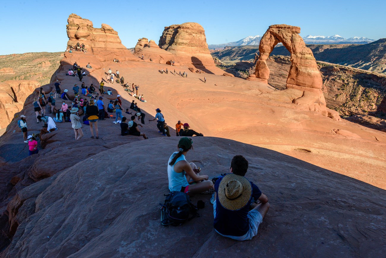 Hikers sit along the edge of a large sandstone bowl as the sun illuminates Delicate arch and several other orange sandstone features.
