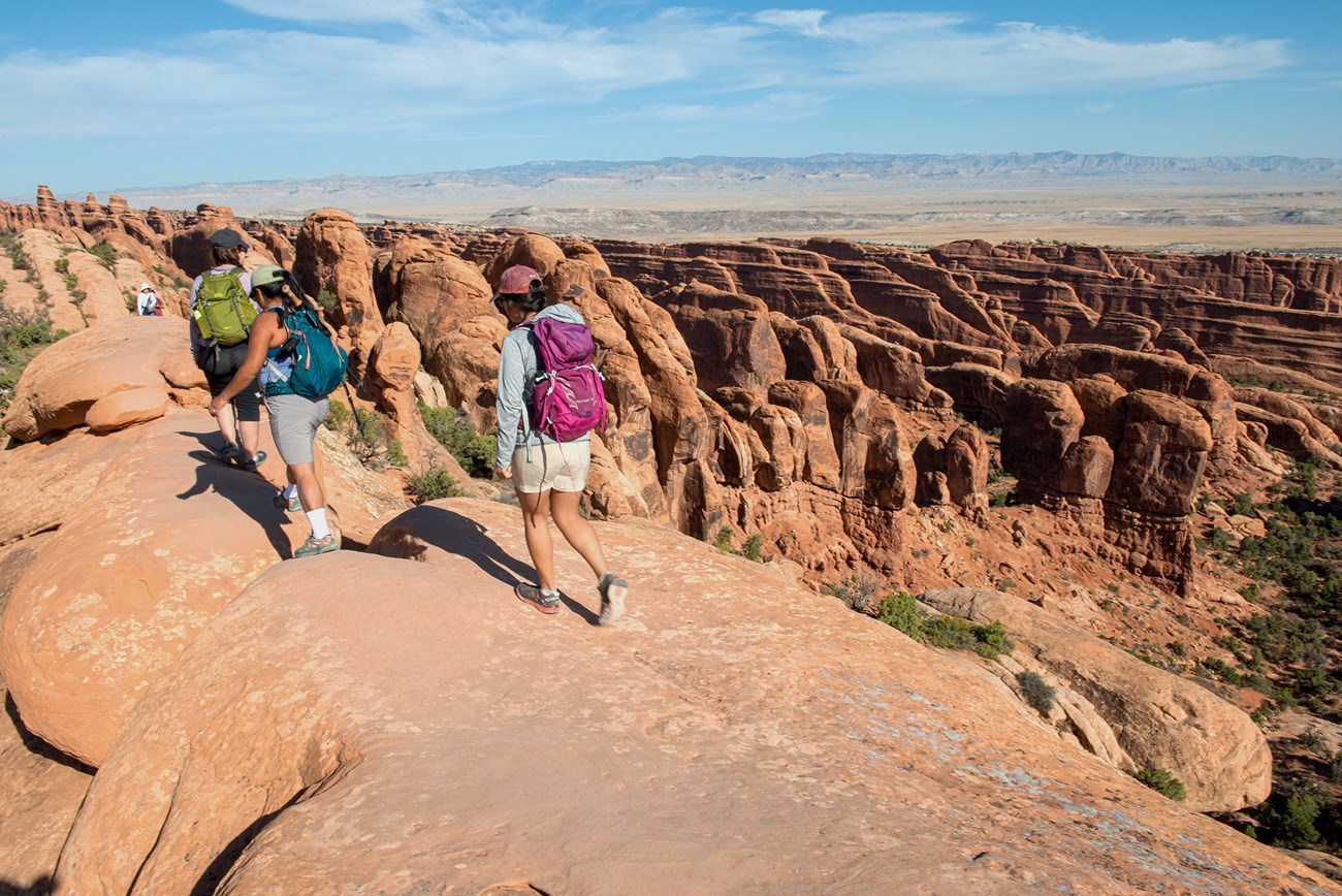 Three hikers in colorful backpacks walk across a long sandstone fin overlooking a series other fins and rock features on a sunny day.