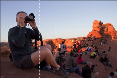 A man points his camera to take a picture. In the background people sit watching the sunlight change on the rock. There is a superimposed grid on the image to break it into three sections. The man with the camera fits in one full section.