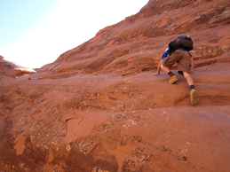 A person in hiking boots and hiking clothes uses their hands to help scramble up a steep red rock face.
