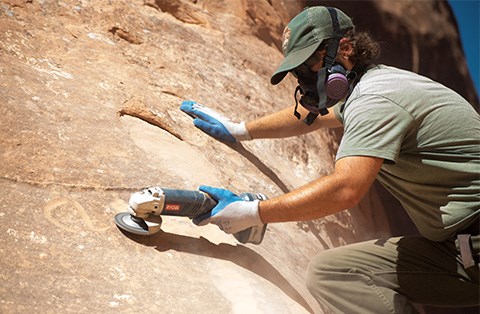A male ranger leans against a rock wall, using a grinding tool to buff out graffiti of someone's name.