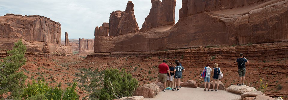 people stand at a paved viewpoint surrounded by high rock walls and looking our across a sandy and green dry landscape.