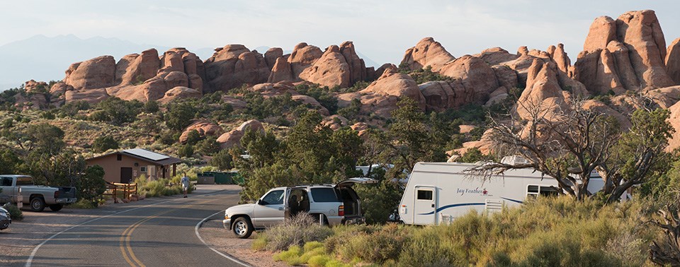 A paved road curves through a campground. Cars and RVs are parked at each campsite. Green vegetation grows around edge of the curving road and large pointed red rocks in the shape of fins are in the background.
