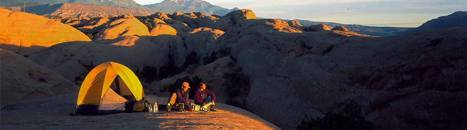 A landscape covered in large smooth rocks with tall green mountains in the distance. Two people sit next on the rocks next to a yellow text cooking a meal. The setting sun is casting a warm glow upon the people, tent, and landscape.