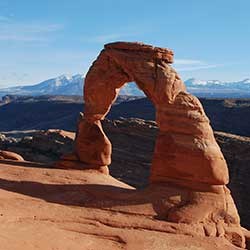 An arch shaped like an upside letter U rises from a sloping red rockface. Through the opening of the arch you can see snow capped mountains and a vast desert landscape. The sky is clear of clouds and light blue.